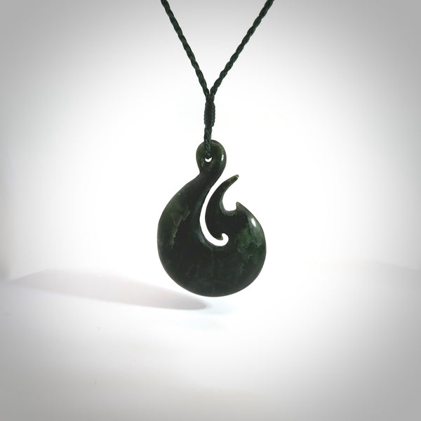 This photo shows a large matau, or hook, pendant carved in deep green New Zealand  Jade. It is a very interesting piece representing a hook. It is strung on a tan cord which is adjustable. The artist who carved this pendant is Ross Crump, a master carver of jade from New Zealand.