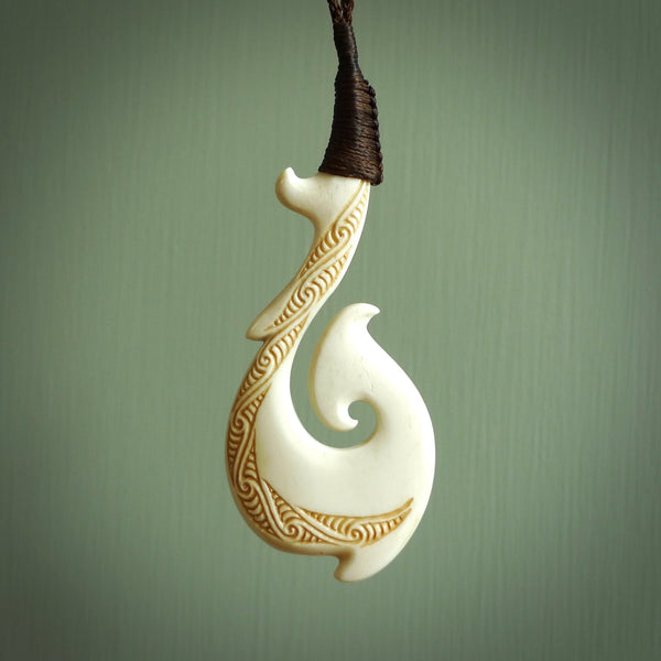 This picture shows a carved hook in bone. The artist has carved traditional decorative kowhaiwhai designs into the body and these run up the insides of the matau. These have specific meanings. It is provided with a hand-plaited brown cord that is length adjustable. Free shipping worldwide.