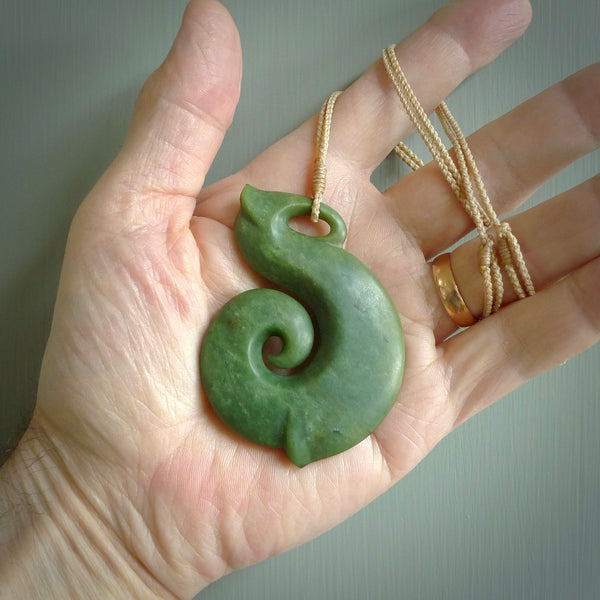 A hand carved hei-matau with koru pendant. This pendant is a green and orange colour and is finished in a polished matte. The cord is a four plait in beige and it is adjustable.