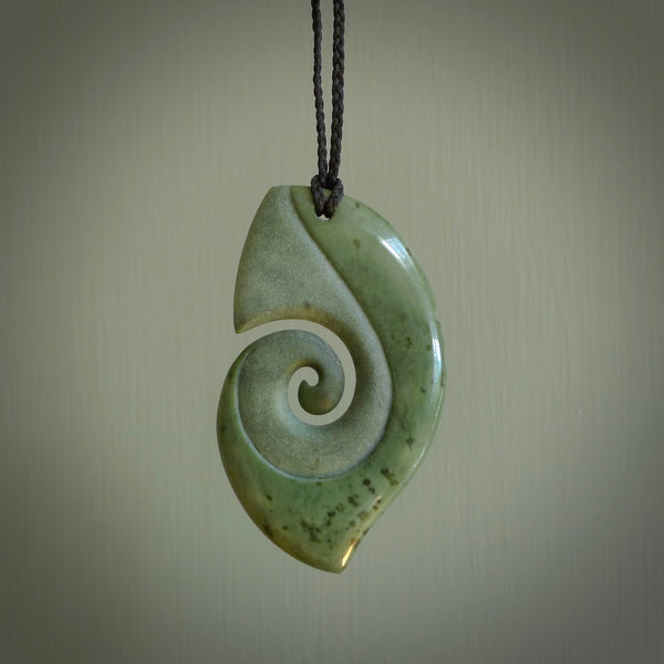 This photo shows a koru with a face carved into the leading edge. The artist, Kerry Thompson, has carved this beautifully and he has polished parts of the carving to a high shine, and other parts he has left in a matte finish. The contrast is beautiful. The pendant is suspended from a plaited black cord which is adjustable.