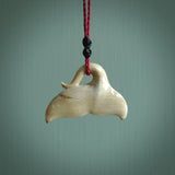 This picture shows a hand carved whale tail pendant. It is a dark, ginger brown colour and is handcarved from Woolly Mammoth tusk. The necklace is made from a braided cord in a pohutukawa red colour.