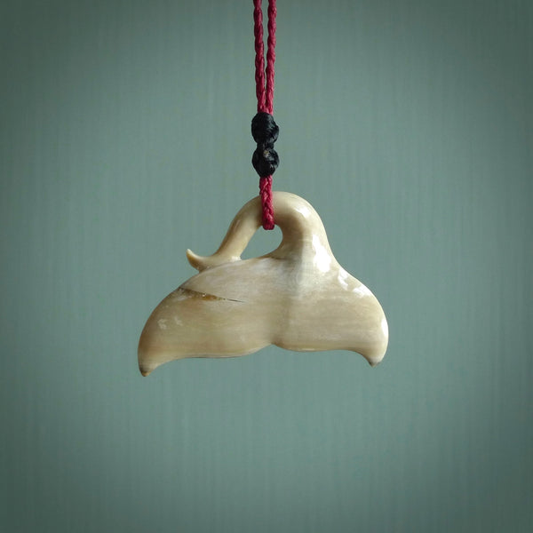 This picture shows a hand carved whale tail pendant. It is a dark, ginger brown colour and is handcarved from Woolly Mammoth tusk. The necklace is made from a braided cord in a pohutukawa red colour.