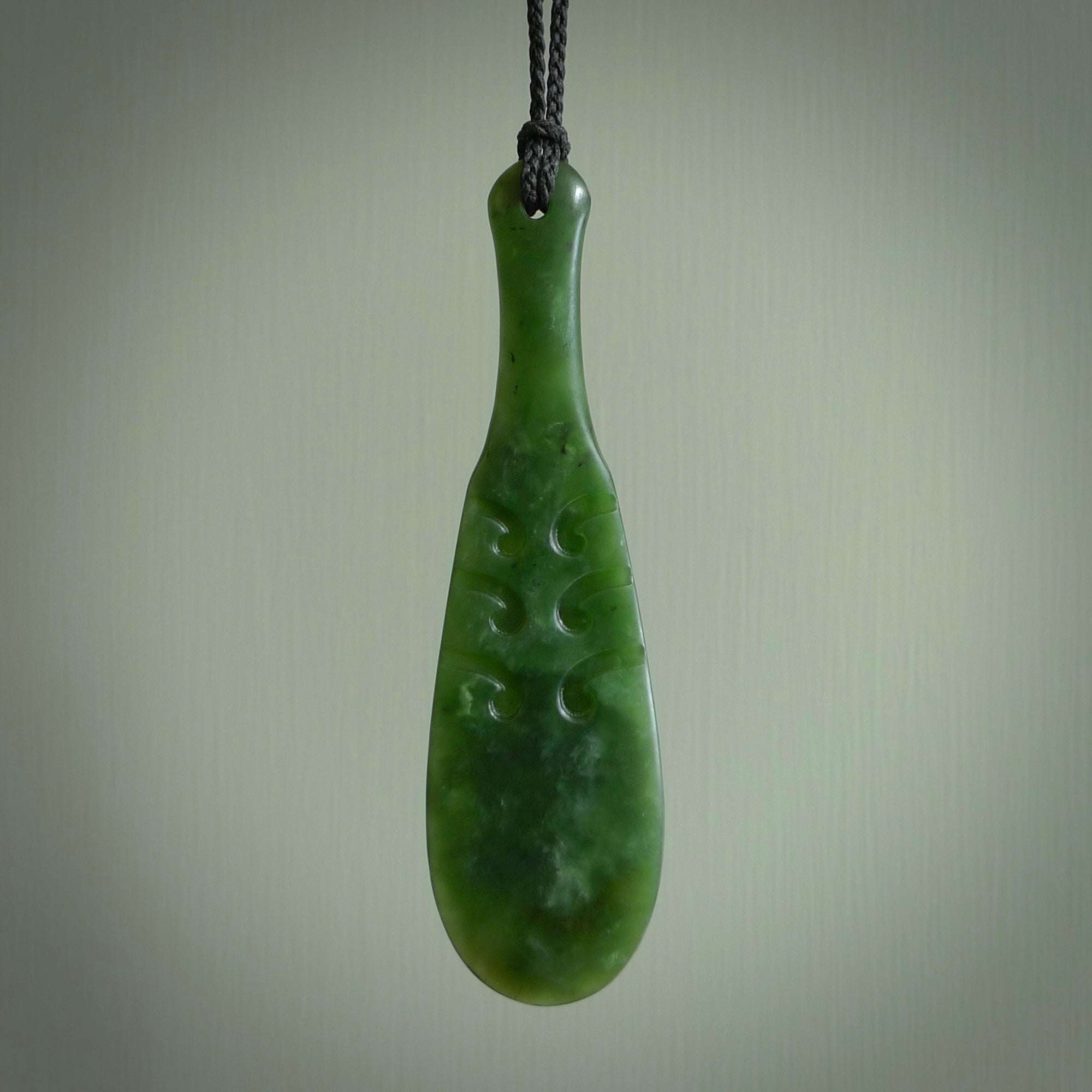 This is a sleek and slender hand carved New Zealand Jade mere pendant. The stone is a very dark green with mottled pale inclusions. The cord is black and adjustable. The pendant has a light polish and just glows. Hand carved by Kerry Thompson. One only pendant.