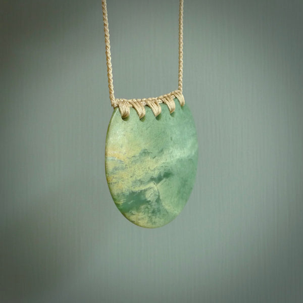 This piece is a large, oval round, disc pendant. It was carved for us by Ric Moor from a lovely milky green piece of New Zealand flower jade. It is suspended on a beige coloured braided cord that is length adjustable.