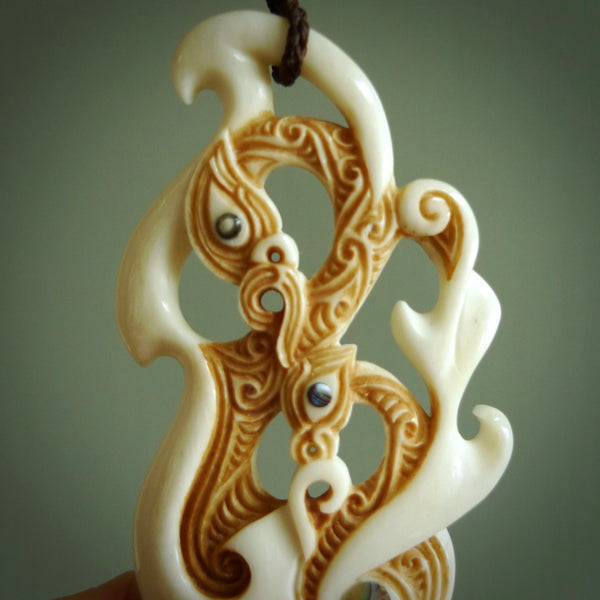 This picture shows a carved double manaia in bone. The artist has carved traditional decorative Koru designs into the face of the body and these run up the sides of the manaia head. These have specific meanings. It is provided with a hand-plaited brown cord that is length adjustable.