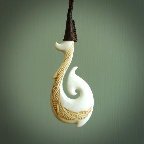 This picture shows a carved hook in bone. The artist has carved traditional decorative kowhaiwhai designs into the body and these run up the insides of the matau. These have specific meanings. It is provided with a hand-plaited brown cord that is length adjustable. Free shipping worldwide.