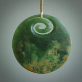 This picture shows a hand carved jade koru pendant. It is a deep green with orange colour and is a wonderful, large sized piece of jewellery. The cord is a four plait beige colour and adjustable so that you can position the pendant where it suits you best. This koru was carved for us by Ric Moor. Delivery is free worldwide.