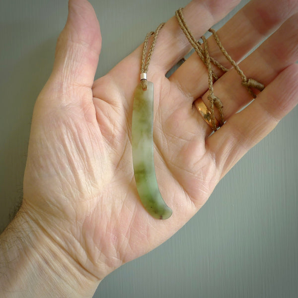 This picture shows a hand carved Spoon River White Jade drop pendant with adjustable cord. The jade is a very light, white green with a shimmer of darker tones in the stone. It is suspended from a tan coloured cord. Delivery is free worldwide.