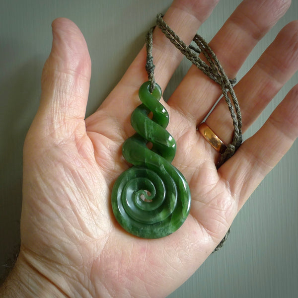 Ross Crump triple twist with koru pendant. Hand carved from rare New Zealand jade this is a beautiful pounamu pendant. The cord is hand plaited in a black colour and is length adjustable. It has a floret in sunrise orange. It is a delicate and very beautiful greenstone pendant. For sale online by NZ Pacific.