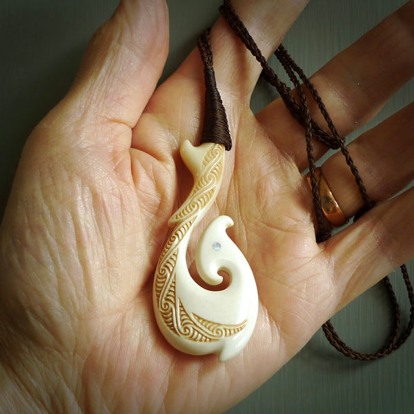 This picture shows a carved hook in bone. The artist has carved traditional decorative kowhaiwhai designs into the body and these run up the insides of the matau. These have specific meanings. It is provided with a hand-plaited brown cord that is length adjustable. Free shipping worldwide.