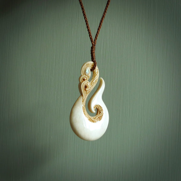 This picture shows a carved manaia in bone. It is a hook shaped pendant pendant that culminates in a fish tail. The artist has carved traditional decorative Koru designs into the side of the body and these run up the sides of the manaia head. These have specific meanings. It is provided with a hand-plaited brown cord that is length adjustable.