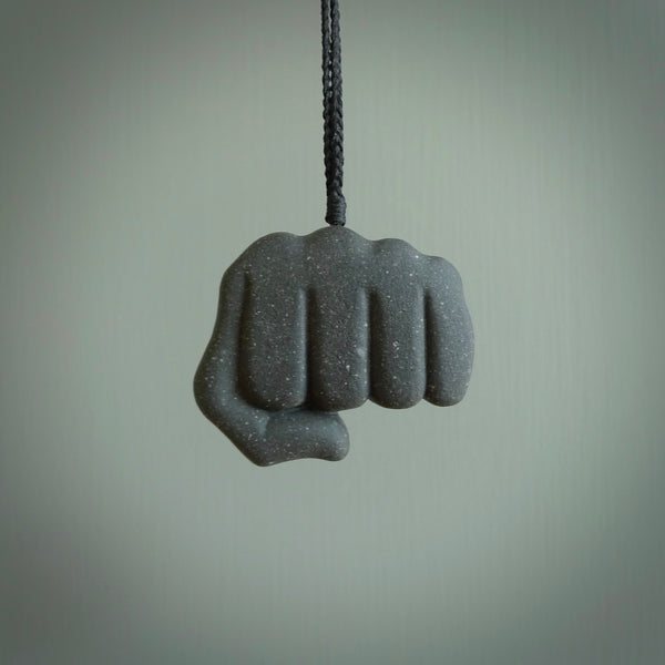 Greywacke Beach Stone fist bump, pendant hand made here in New Zealand. Hand carved by Rhys Hall for NZ Pacific. Handmade jewellery for sale online. The cord is black and has a loop and pebble toggle closure. Unique necklace for men and women. fist pump necklace hand made from New Zealand Greywacke Beach Stone.