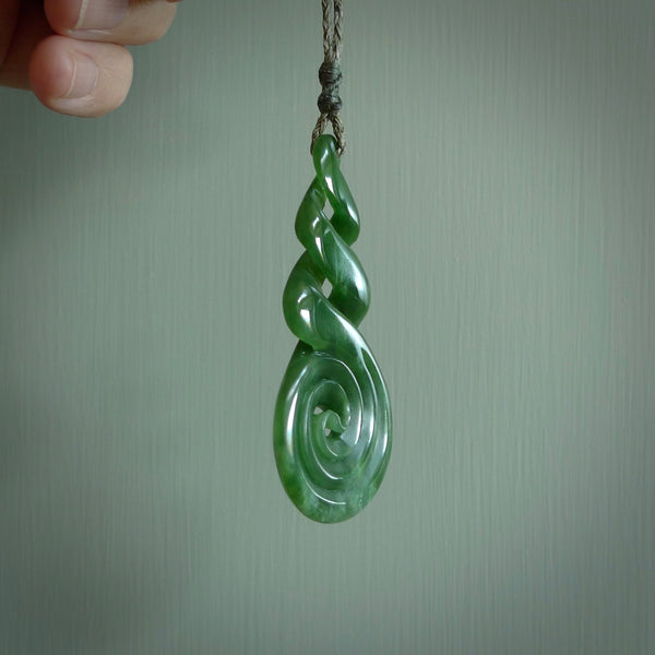 Ross Crump triple twist with koru pendant. Hand carved from rare New Zealand jade this is a beautiful pounamu pendant. The cord is hand plaited in a black colour and is length adjustable. It has a floret in sunrise orange. It is a delicate and very beautiful greenstone pendant. For sale online by NZ Pacific.