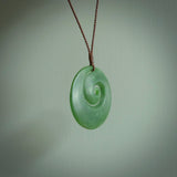This picture shows a large hand carved jade koru pendant. It is a darker green colour which is quite translucent where the jade is thin. Although a large piece, it is a wonderful, delicate piece of jewellery. The cord is hand plaited and adjustable so that you can position the pendant where it suits you best. This koru was carved for us by Ric Moor. Delivery is free worldwide.