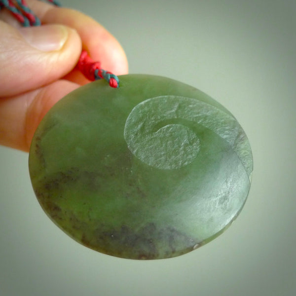 Hand carved jade disc pendant. One side has a single koru etched into the face. This is a subtle and beautiful design. The other side is plain. The cord is a Cobalt Blue/Juicy Red colour and is adjustable. The piece was carved for NZ Pacific by Rueben Tipene.
