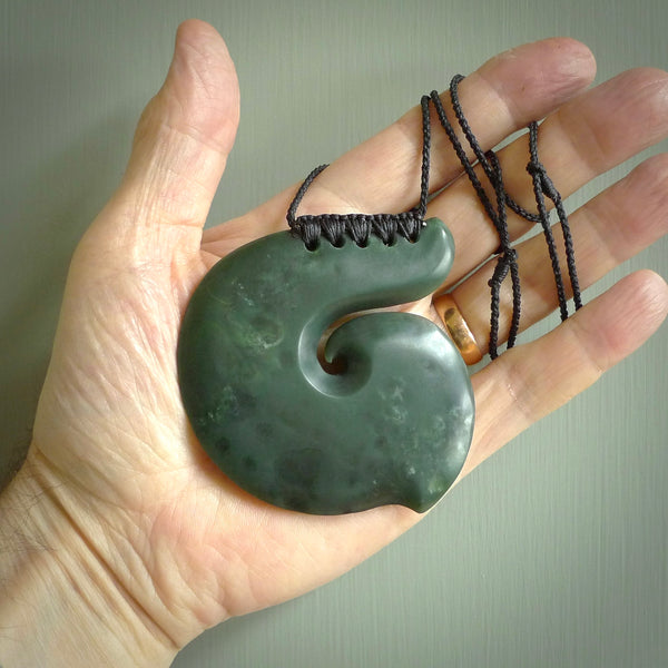 This picture shows a hand carved, large, jade koru pendant. It is a deep green colour and is a wonderful, large sized piece of jewellery. The cord is a four plait beige colour and adjustable so that you can position the pendant where it suits you best. This koru was carved for us by Ric Moor. Delivery is free worldwide.