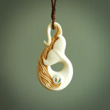 This picture shows a carved whale tail in bone. It is a hook shaped pendant pendant that culminates in a fish tail. The artist has carved traditional decorative Koru designs into the body and these run up the inner sides of the hook. These have specific meanings. It is provided with a plaited brown coloured cord that is length adjustable.