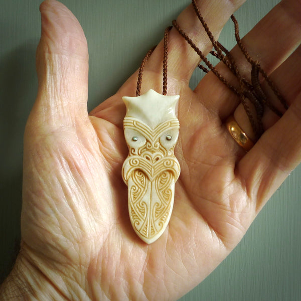 This is a large carved Koruru pendant. It is made from two materials - New Zealand Paua Shell and Bone. The face of the Koruru is carved with a traditional design in the front. It has a protruding tongue which is also carved and the back face has traditional carved designs. This is a large and very traditional pendant that is a collectors piece. This piece is provided on a brown adjustable cord.