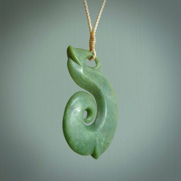 A hand carved hei-matau with koru pendant. This pendant is a green and orange colour and is finished in a polished matte. The cord is a four plait in beige and it is adjustable.