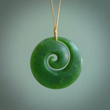 This picture shows a hand carved jade koru pendant. It is a deep green with orange colour and is a wonderful, large sized piece of jewellery. The cord is a four plait beige colour and adjustable so that you can position the pendant where it suits you best. This koru was carved for us by Ric Moor. Delivery is free worldwide.