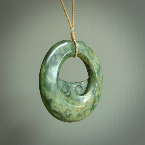 This piece is a large sized, oval round, disc pendant with double koru heart. It was carved for us by Ric Moor from a lovely deep and milky green piece of New Zealand flower jade. It is suspended on a beige  coloured braided cord that is length adjustable.