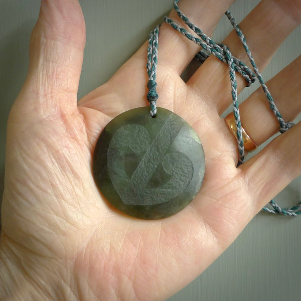 Hand carved jade disc pendant. One side has a double koru etched into the face. This is a subtle and beautiful design. The other side is plain. The cord is a tan colour and is adjustable. The piece was carved for NZ Pacific by Rueben Tipene.