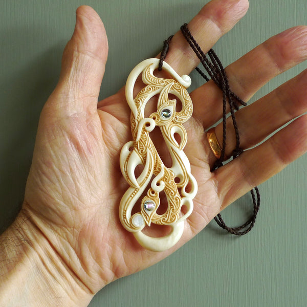 This picture shows a carved double manaia in bone. The artist has carved traditional decorative Koru designs into the face of the body and these run up the sides of the manaia head. These have specific meanings. It is provided with a hand-plaited brown cord that is length adjustable.