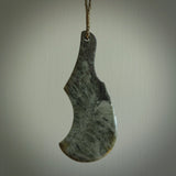This is a sleek and slender hand carved New Zealand Jade mere pendant. The stone is a very dark green with mottled pale inclusions. The cord is tan and adjustable. The pendant has a light polish and just glows.