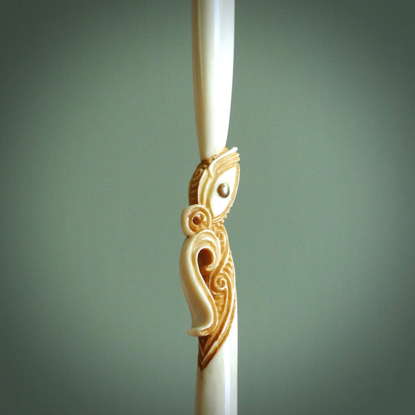 This picture shows a carved aurei, cloak pin in bone. It is a large pendant that is sure to catch attention. The artist has carved traditional decorative Koru designs into the manaia face. These have specific meanings. It is provided with a plaited brown coloured cord that is length adjustable.