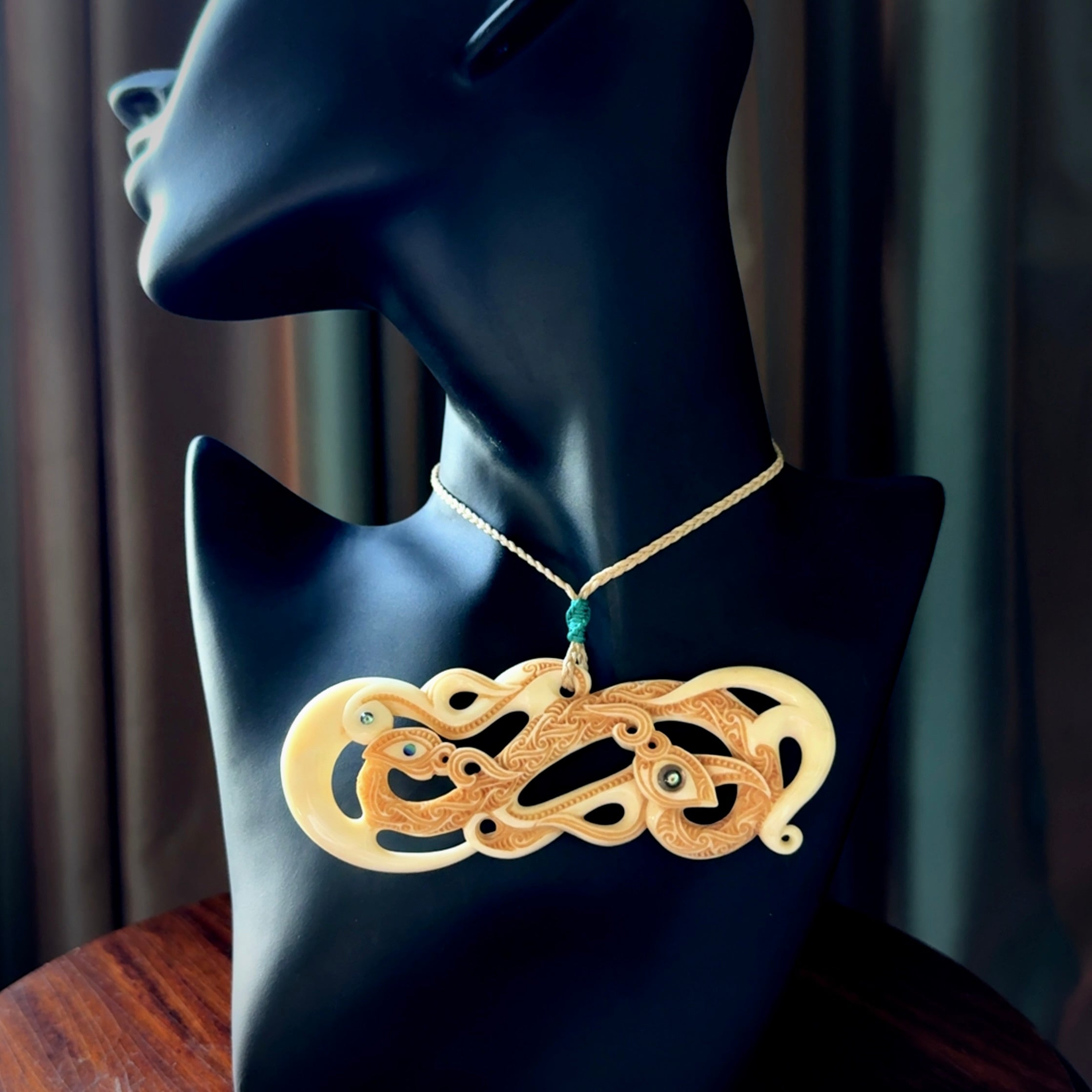 This picture shows a carved double manaia in bone. The artist has carved traditional decorative Koru designs into the face of the body and these run up the sides of the manaia head. These have specific meanings. It is provided with a hand-plaited oat cord that is length adjustable.