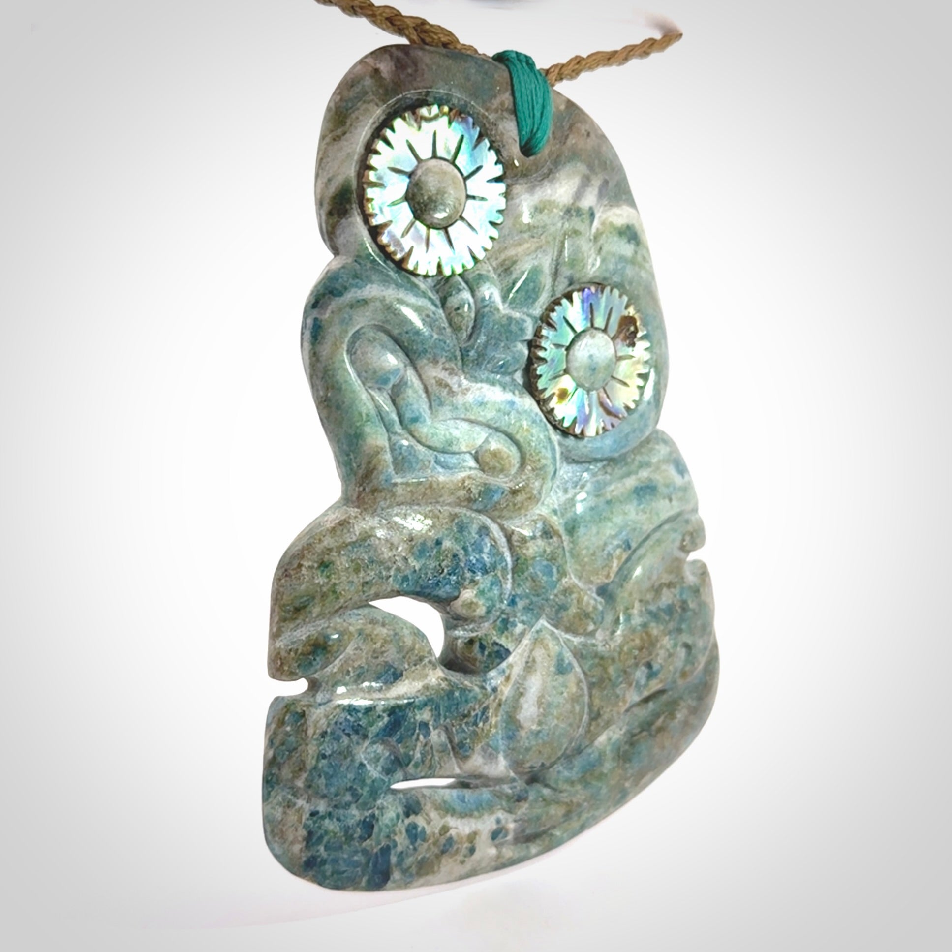 This photo shows a large hand carved Aotea stone Tiki pendant. It is a very light blue colour with pāua shell insert eyes. The cord is a thick camel colour and is length adjustable