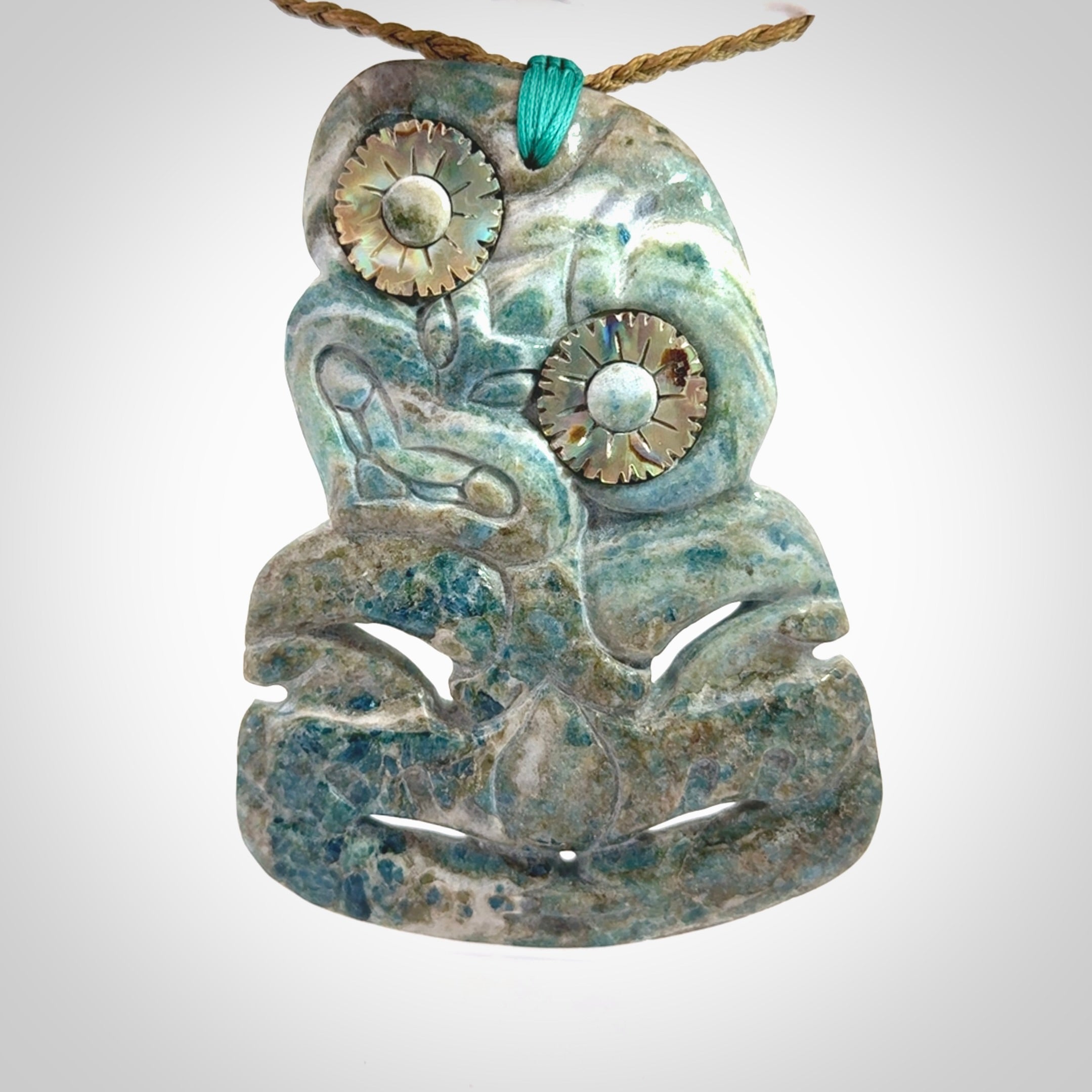 This photo shows a large hand carved Aotea stone Tiki pendant. It is a very light blue colour with pāua shell insert eyes. The cord is a thick camel colour and is length adjustable