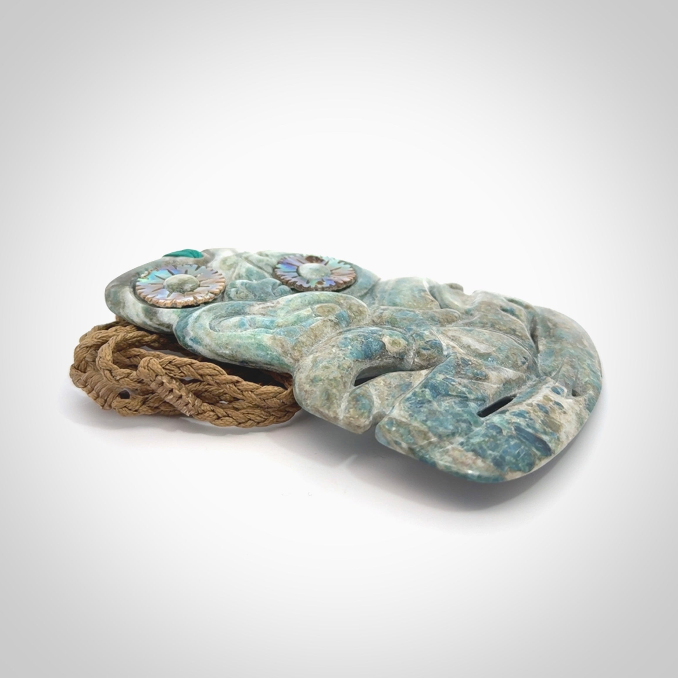 This photo shows a large hand carved Aotea stone Tiki pendant. It is a very light blue colour with pāua shell insert eyes. The cord is a thick camel colour and is length adjustable