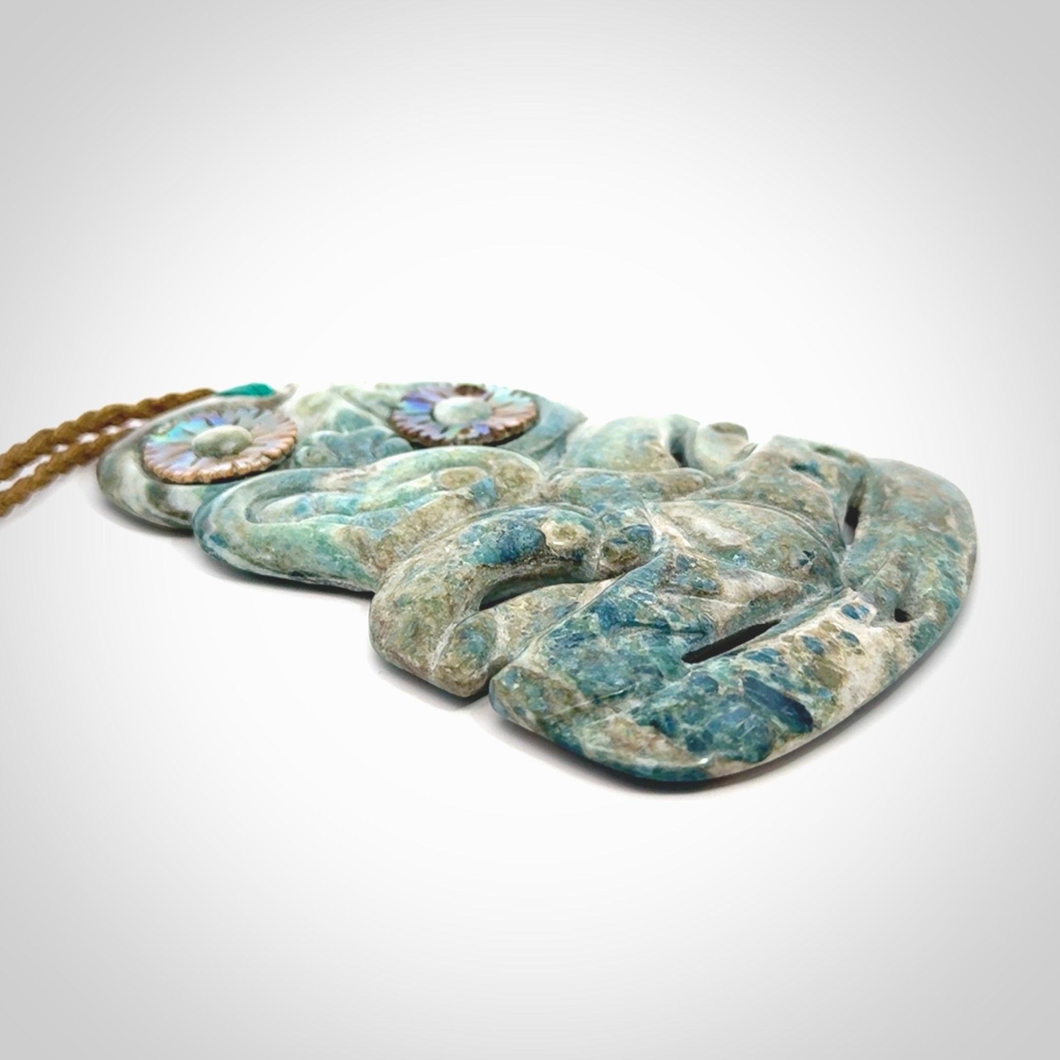 This photo shows a large hand carved Aotea stone Tiki pendant. It is a very light blue colour with pāua shell insert eyes. The cord is a thick camel colour and is length adjustable