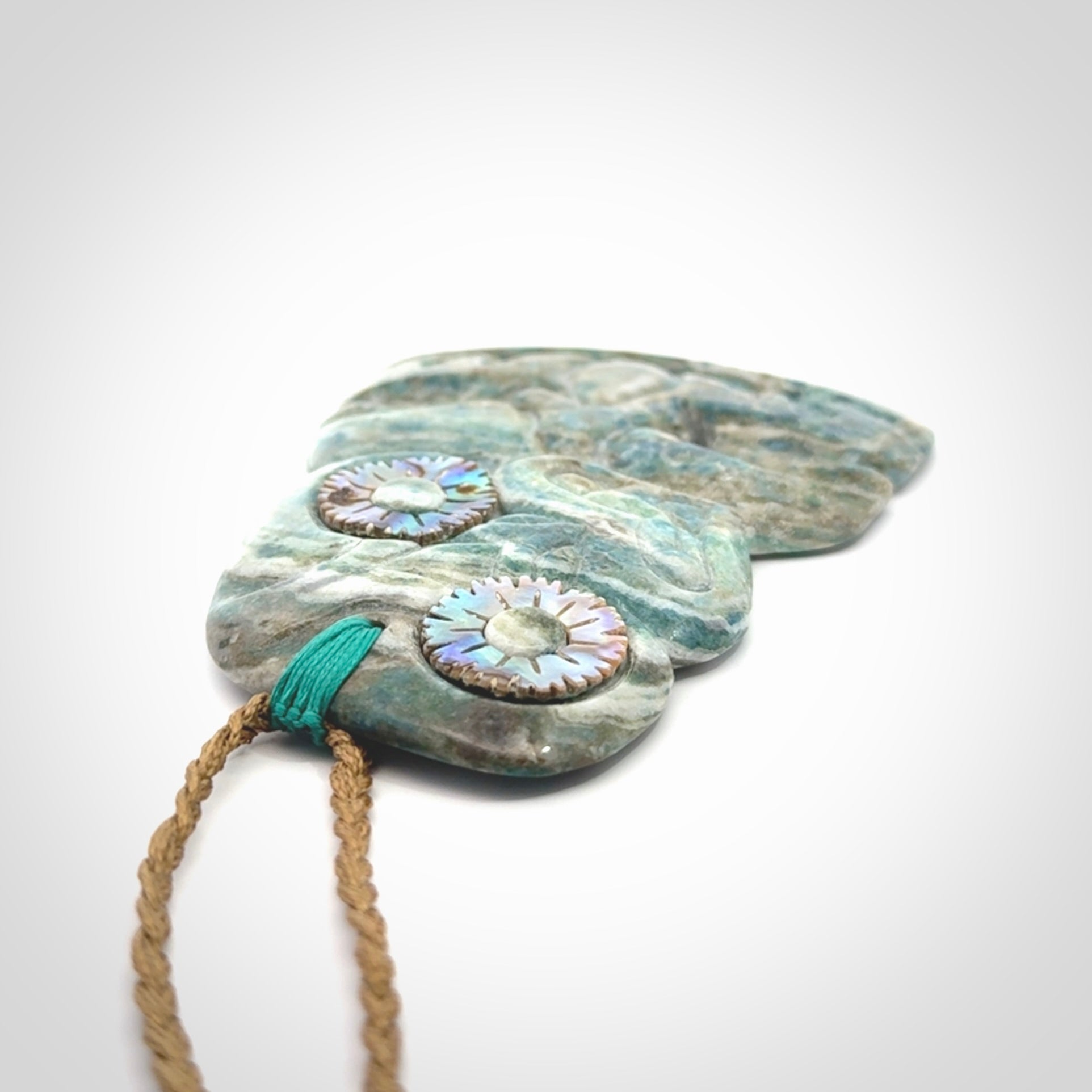 This photo shows a large hand carved Aotea stone Tiki pendant. It is a very light blue colour with pāua shell insert eyes. The cord is a thick camel colour and is length adjustable