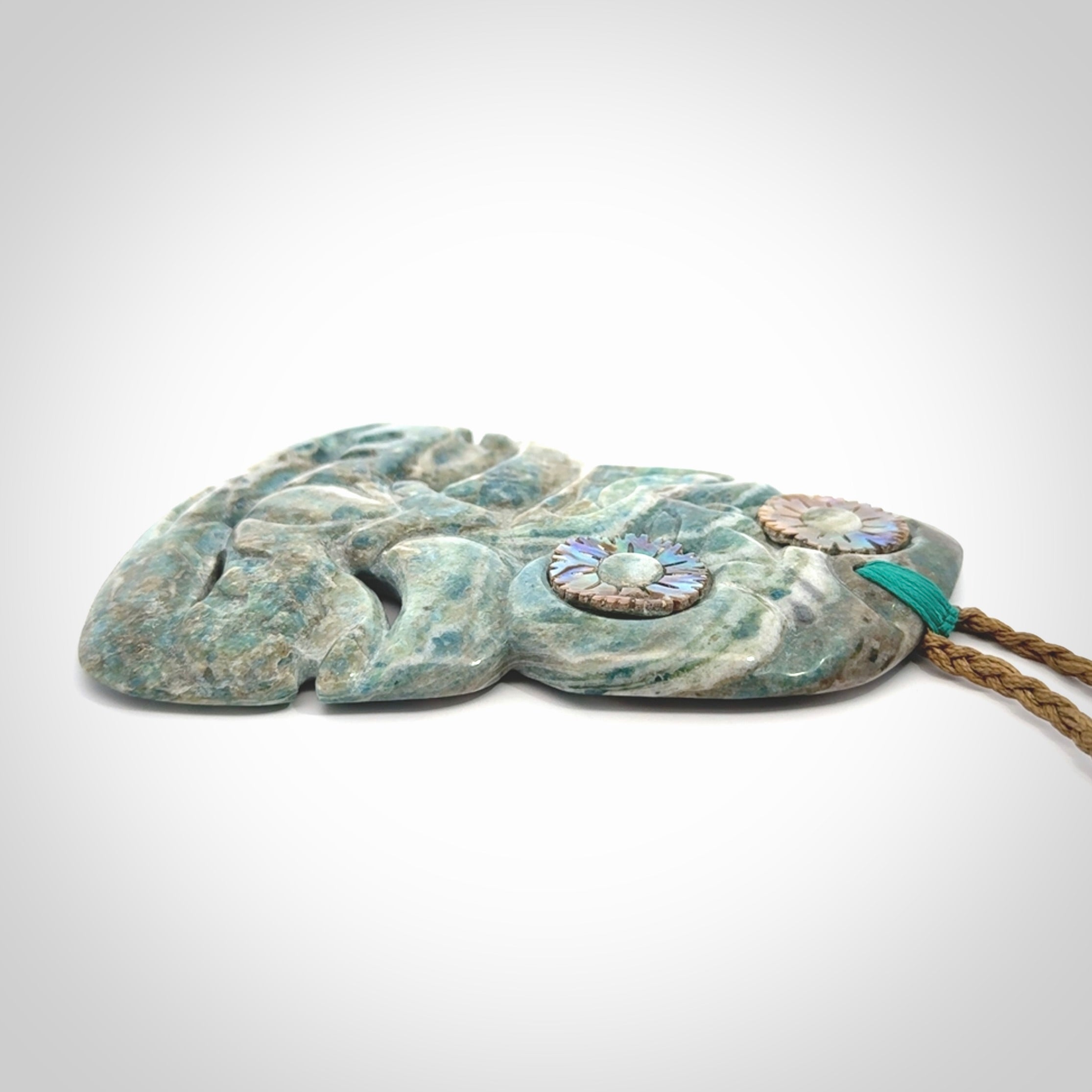 This photo shows a large hand carved Aotea stone Tiki pendant. It is a very light blue colour with pāua shell insert eyes. The cord is a thick camel colour and is length adjustable