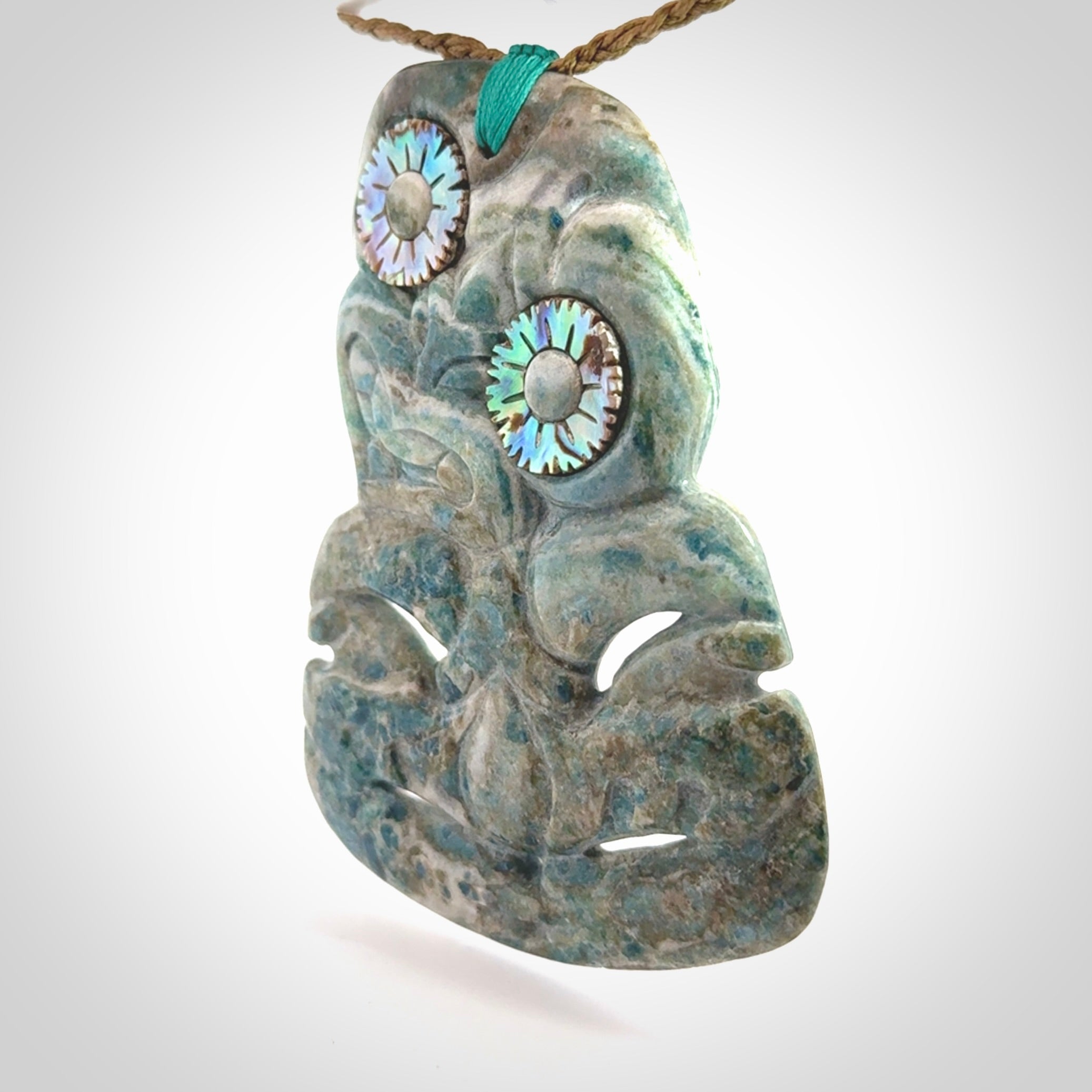 This photo shows a large hand carved Aotea stone Tiki pendant. It is a very light blue colour with pāua shell insert eyes. The cord is a thick camel colour and is length adjustable
