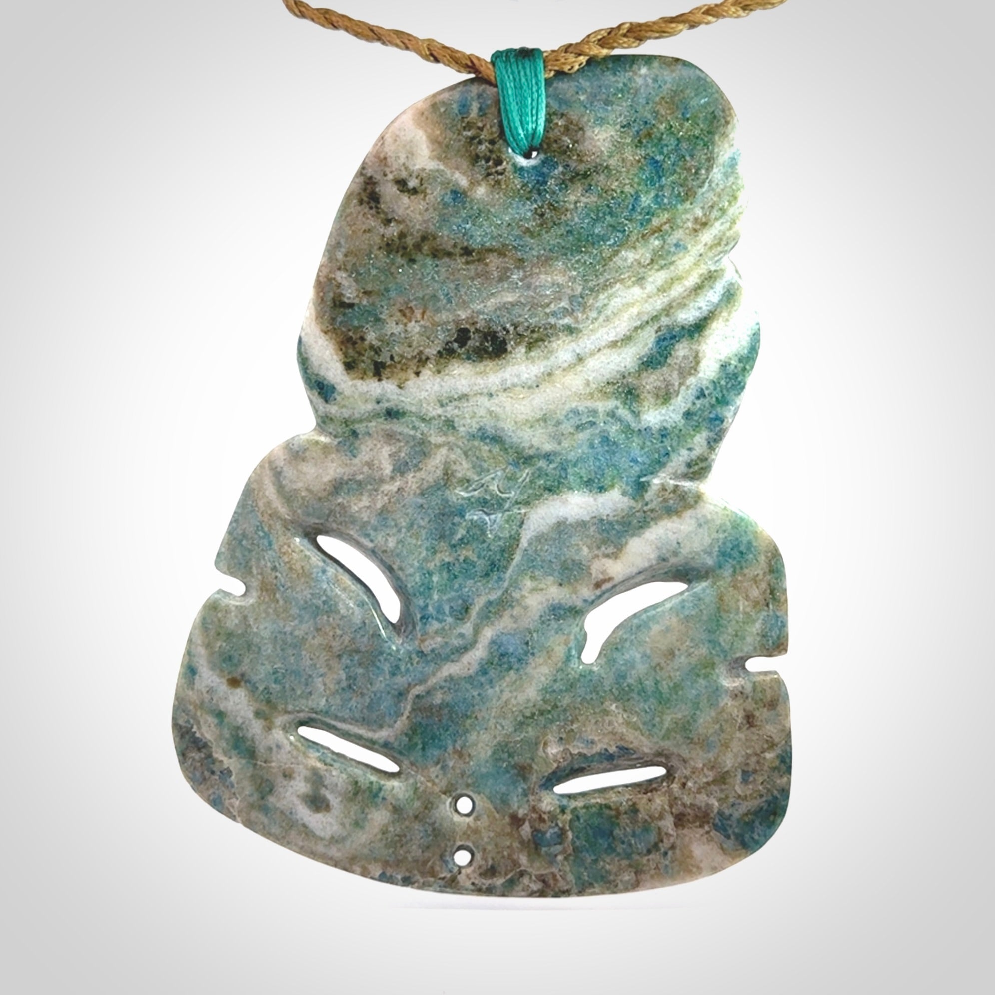 This photo shows a large hand carved Aotea stone Tiki pendant. It is a very light blue colour with pāua shell insert eyes. The cord is a thick camel colour and is length adjustable