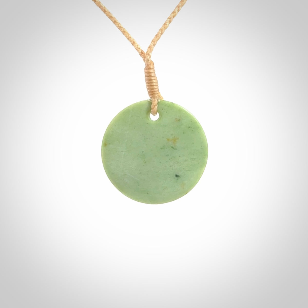 This piece is a small oval round, disc pendant carved in New Zealand Jade. It is suspended on a beige coloured braided cord that is length adjustable.