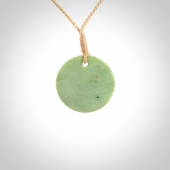 This piece is a small oval round, disc pendant carved in New Zealand Jade. It is suspended on a beige coloured braided cord that is length adjustable.