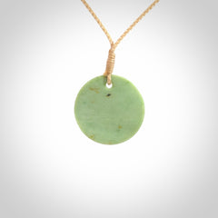 This piece is a small oval round, disc pendant carved in New Zealand Jade. It is suspended on a beige coloured braided cord that is length adjustable.