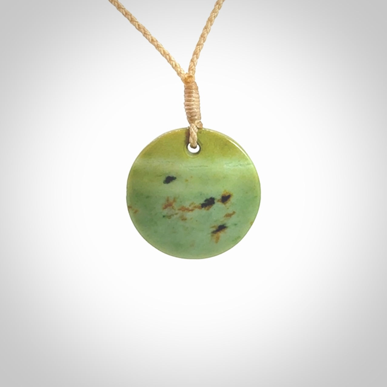 This piece is a small oval round, disc pendant carved in New Zealand Flower Jade. It is suspended on a beige coloured braided cord that is length adjustable.