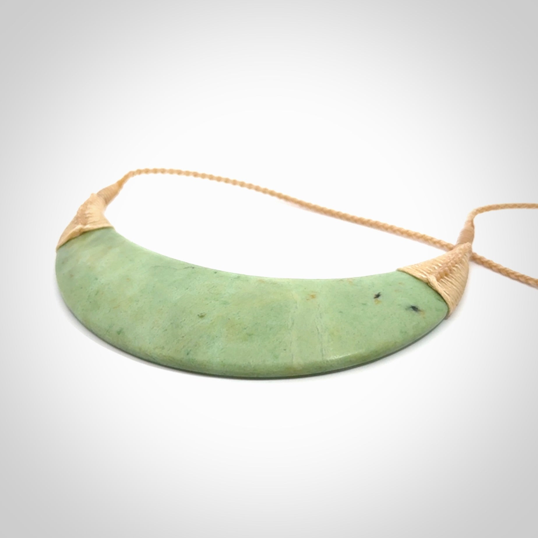 This picture shows a hand carved New Zealand jade contemporary half-shield pendant. It is a light green colour and is a wonderful, large piece of jewellery. The cord is hand plaited and adjustable so that you can position the pendant where it suits you best. This piece was carved for us by Donna Summers.