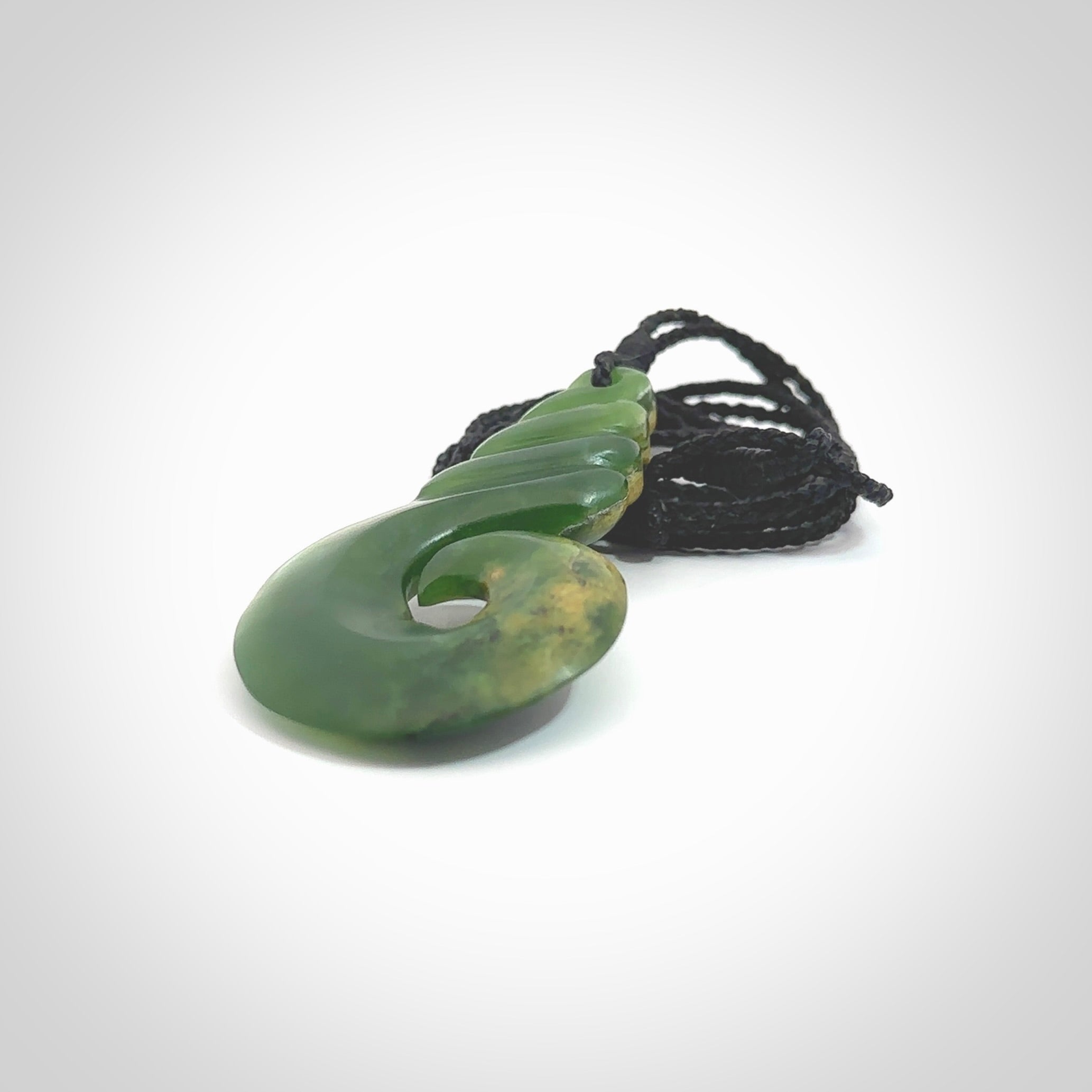 This picture shows a hook shaped pendant, hand carved from New Zealand flower jade. The top of the hook, the shaft, is a twist shape. We will provide this with an adjustable plaited cord. The cord is a adjustable and is a black colour. Twist and koru designs are also displayed in this stunning New Zealand Marsden Flower Jade pendant. Hand made by Ana Krakosky.