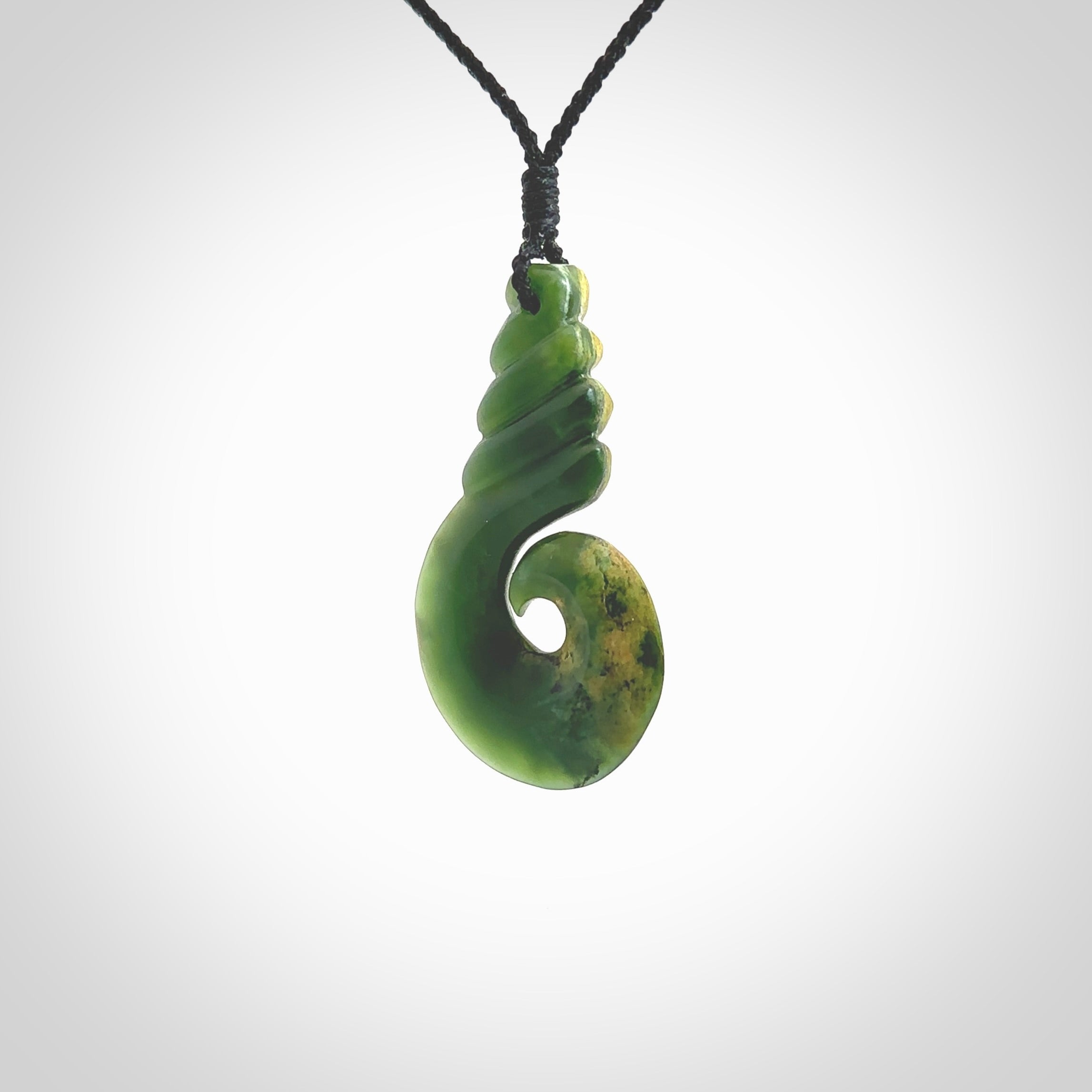This picture shows a hook shaped pendant, hand carved from New Zealand flower jade. The top of the hook, the shaft, is a twist shape. We will provide this with an adjustable plaited cord. The cord is a adjustable and is a black colour. Twist and koru designs are also displayed in this stunning New Zealand Marsden Flower Jade pendant. Hand made by Ana Krakosky.