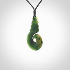 This picture shows a hook shaped pendant, hand carved from New Zealand flower jade. The top of the hook, the shaft, is a twist shape. We will provide this with an adjustable plaited cord. The cord is a adjustable and is a black colour. Twist and koru designs are also displayed in this stunning New Zealand Marsden Flower Jade pendant. Hand made by Ana Krakosky.