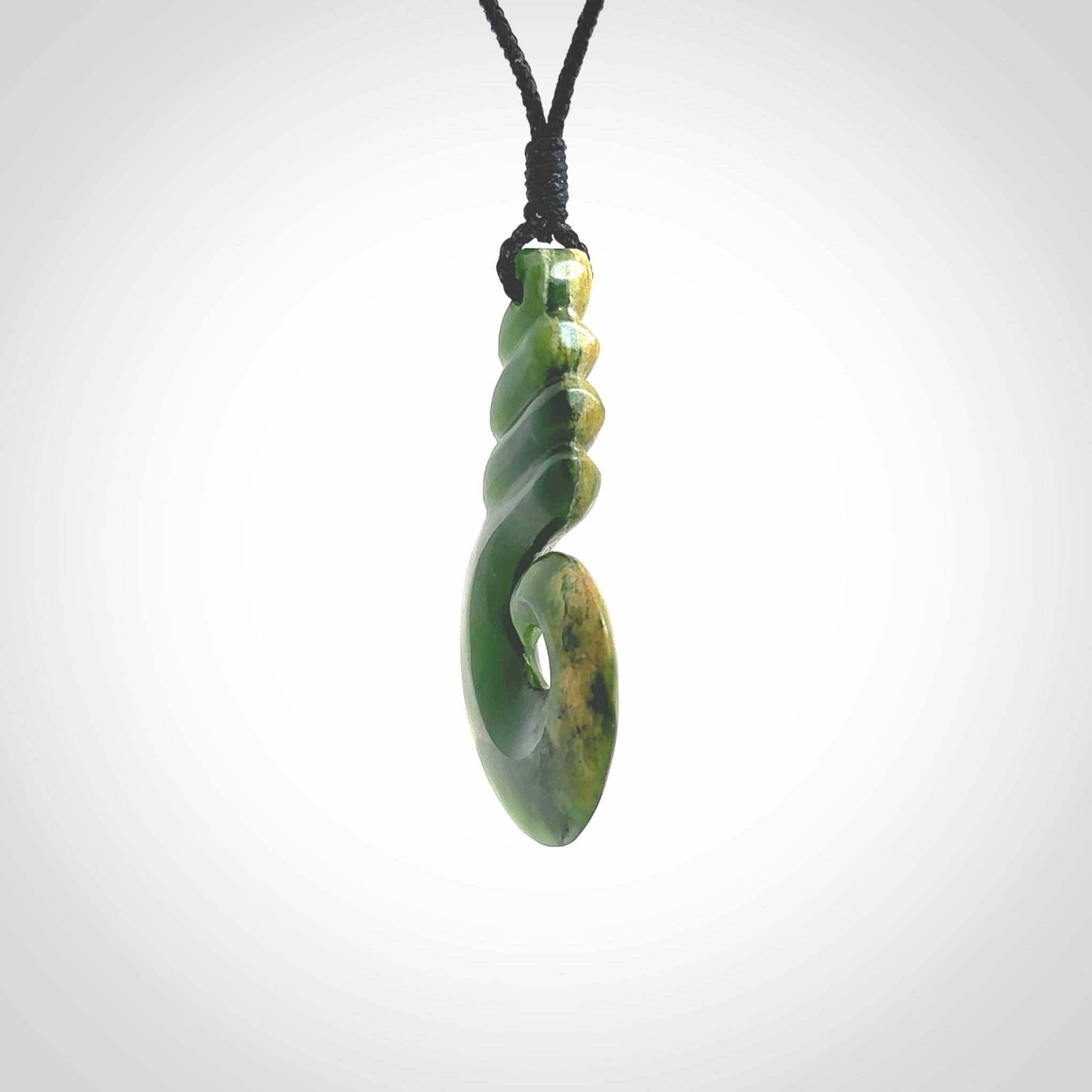 This picture shows a hook shaped pendant, hand carved from New Zealand flower jade. The top of the hook, the shaft, is a twist shape. We will provide this with an adjustable plaited cord. The cord is a adjustable and is a black colour. Twist and koru designs are also displayed in this stunning New Zealand Marsden Flower Jade pendant. Hand made by Ana Krakosky.