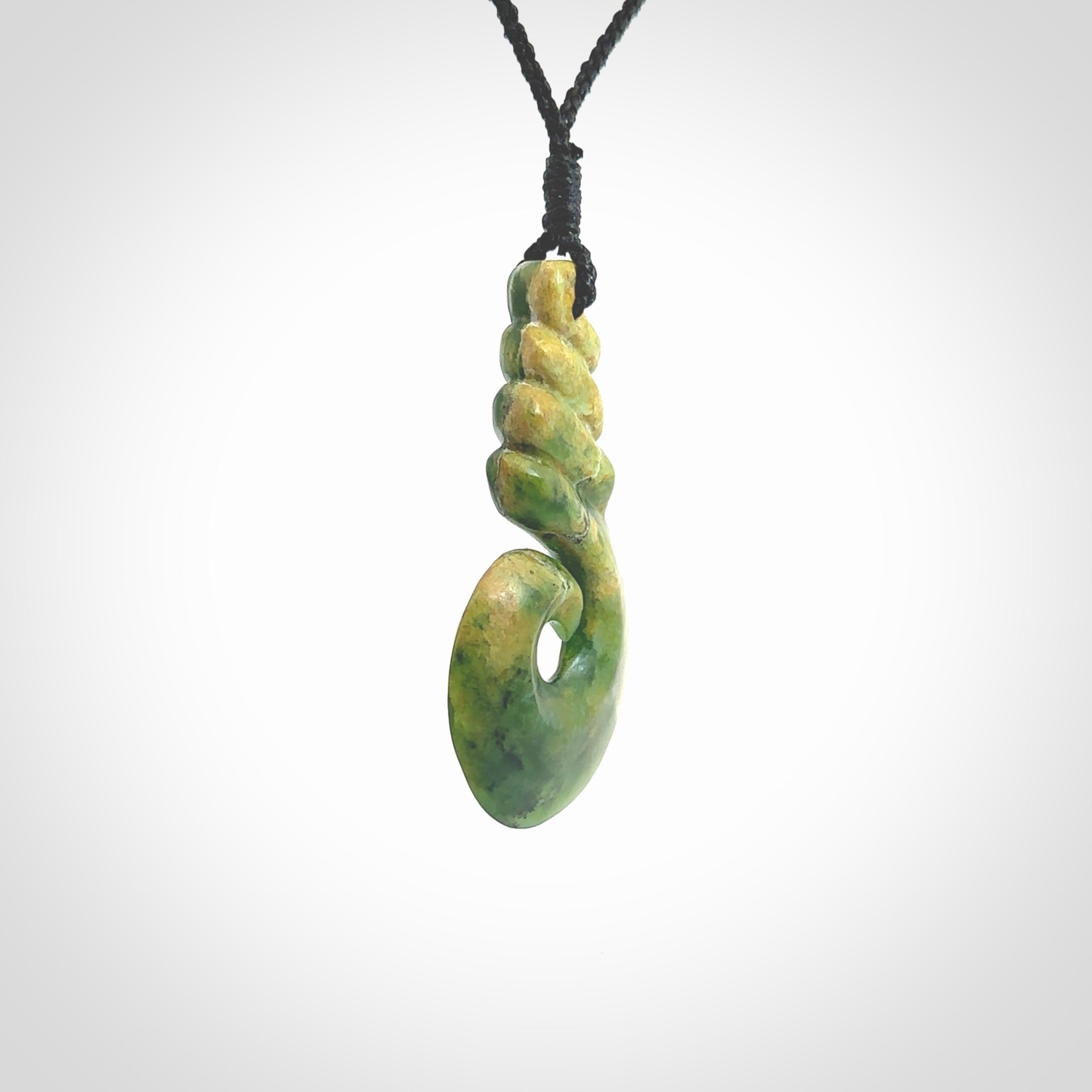 This picture shows a hook shaped pendant, hand carved from New Zealand flower jade. The top of the hook, the shaft, is a twist shape. We will provide this with an adjustable plaited cord. The cord is a adjustable and is a black colour. Twist and koru designs are also displayed in this stunning New Zealand Marsden Flower Jade pendant. Hand made by Ana Krakosky.