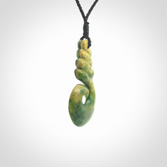 This picture shows a hook shaped pendant, hand carved from New Zealand flower jade. The top of the hook, the shaft, is a twist shape. We will provide this with an adjustable plaited cord. The cord is a adjustable and is a black colour. Twist and koru designs are also displayed in this stunning New Zealand Marsden Flower Jade pendant. Hand made by Ana Krakosky.