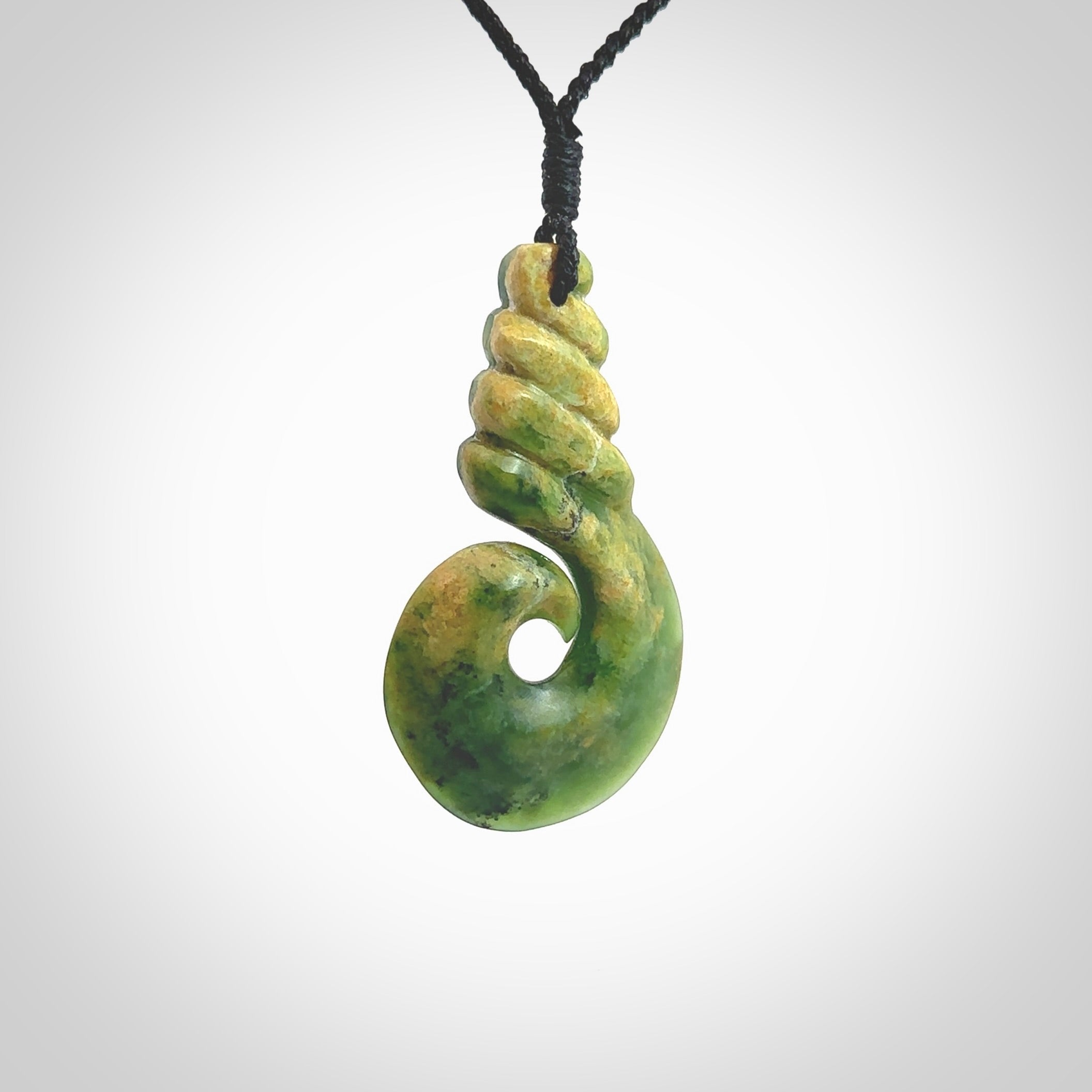 This picture shows a hook shaped pendant, hand carved from New Zealand flower jade. The top of the hook, the shaft, is a twist shape. We will provide this with an adjustable plaited cord. The cord is a adjustable and is a black colour. Twist and koru designs are also displayed in this stunning New Zealand Marsden Flower Jade pendant. Hand made by Ana Krakosky.